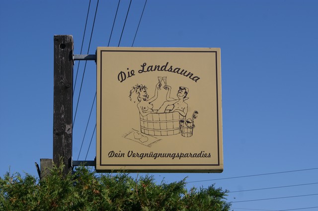 Schild an einem Mast. Text: Die Landsauna, Dein Vergnügungsparadies. Zeichnung von zwei Personen in einem Zuber.