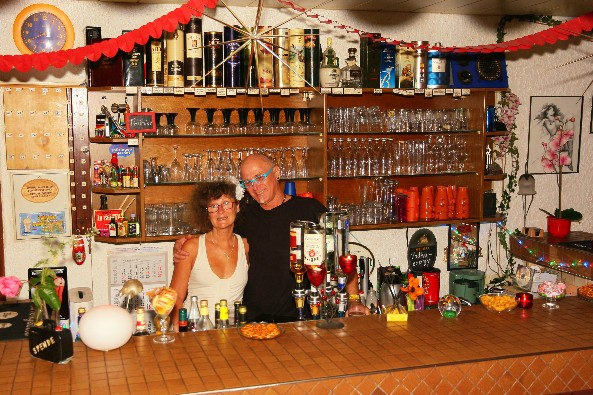 Zwei Personen hinter einer Bar, Regale mit Flaschen und Gläsern.